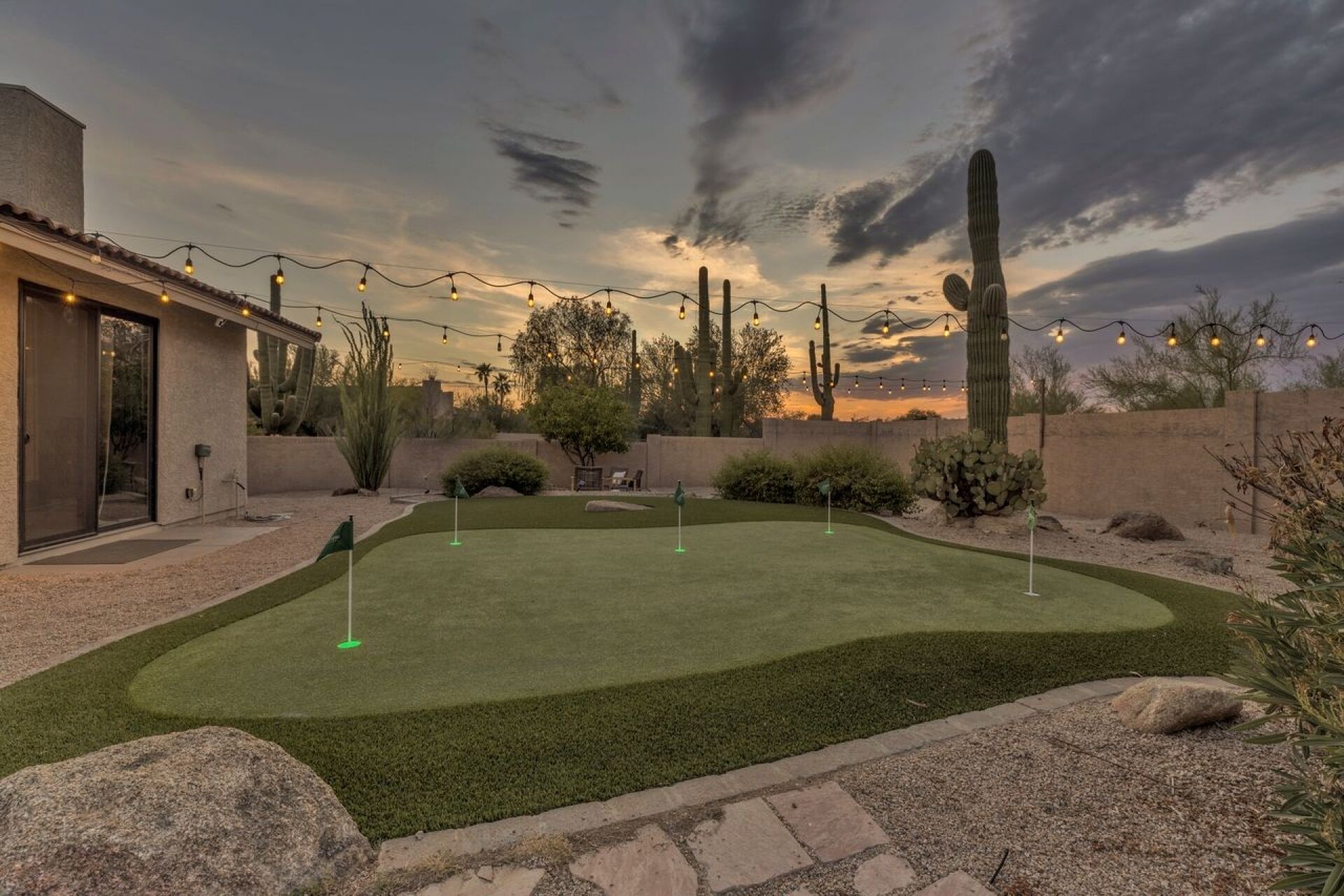 A well-maintained artificial grass golf course with a house and cacti in the background.