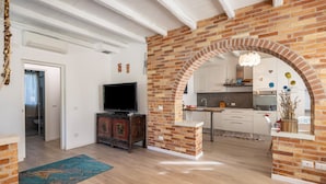 Una sala de estar moderna con televisor, suelo de madera y un arco de ladrillo que conduce a una cocina.