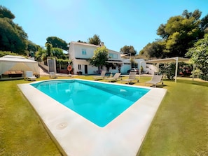 A rectangular swimming pool with a white deck, surrounded by a well-maintained lawn and a white building with a tiled roof in the background.