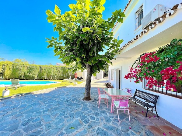A courtyard with a swimming pool, a tree, pink chairs, and a bench.