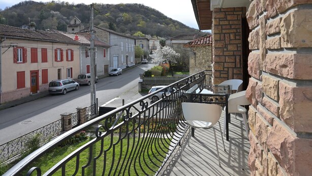 A balcony with a stone wall, a metal railing, and a view of a street with parked cars and buildings.