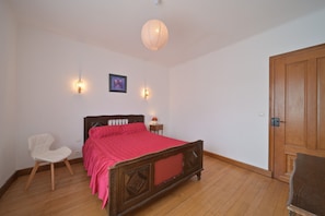 Une chambre à coucher avec un lit en bois, une chaise, une table de chevet, une lampe fixée au mur et une porte.