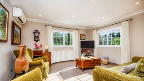 A living room with a sofa, a coffee table, a TV, a clock, and a window. - Villa Puerto Apartment with Mountain Views, Pool, and Wi-Fi (Sóller)