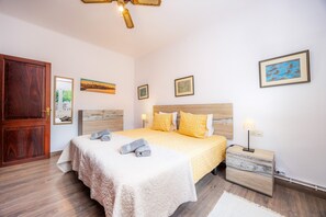 Quarto com porta de madeira, cama com colcha bege, duas almofadas, mesa de cabeceira com abajur e quadros nas paredes.