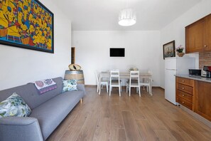 A living room with a sofa, a dining table with chairs, a kitchen area with cabinets, a TV mounted on the wall, and a large painting on the wall. - Casa Arinto – Spacious, Peaceful Retreat Among Vineyards with Lounge & Shared Pool (Palmela)