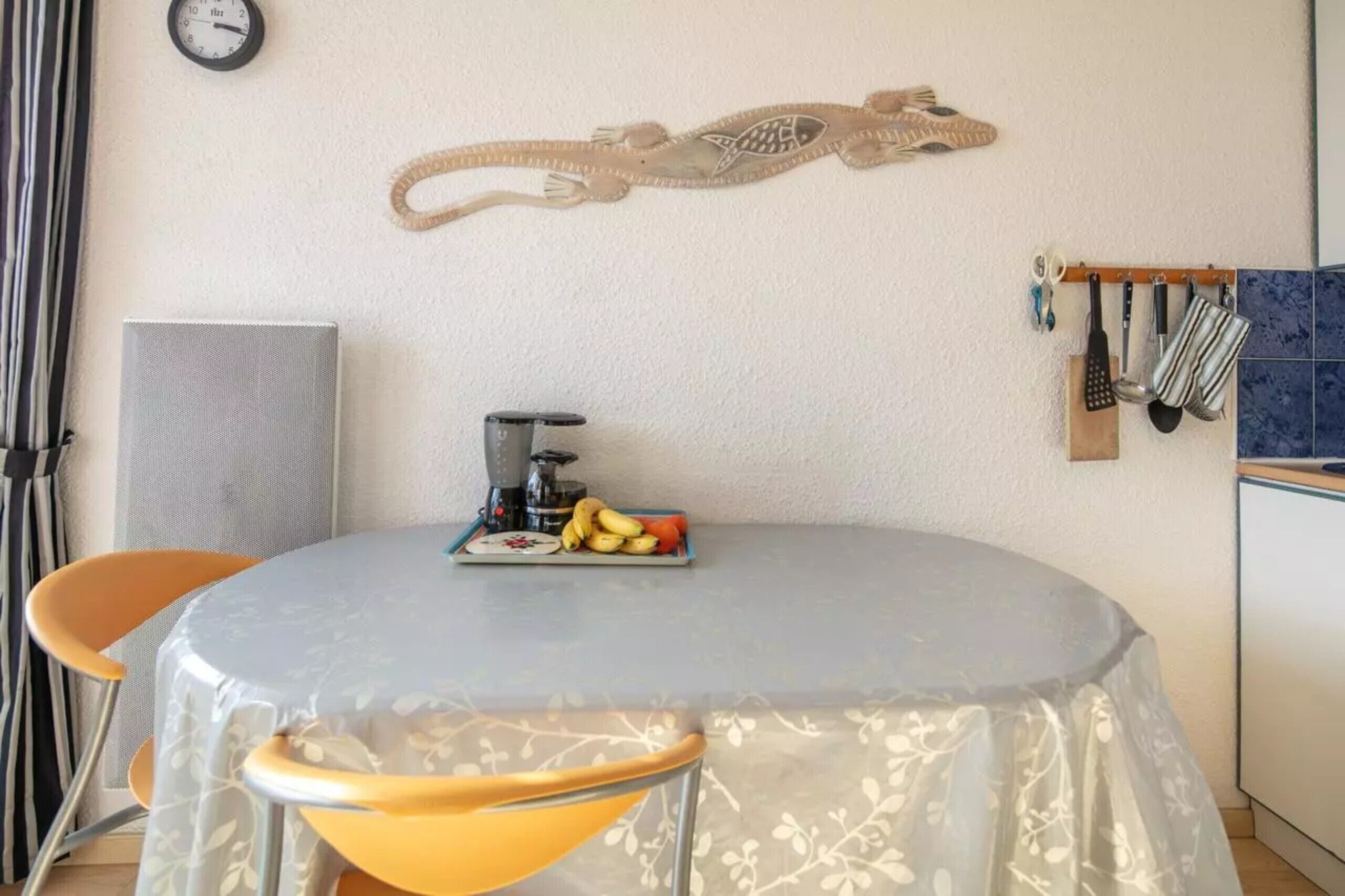 Une table ronde recouverte d’une nappe à motifs floraux, une machine à café et une assiette de fruits.