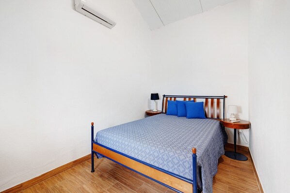 A bedroom with a bed, a nightstand, a lamp, and a wall-mounted air conditioner.
