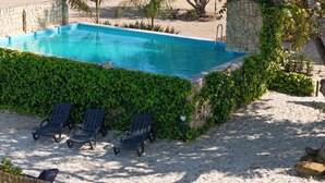 Une piscine bordée d’un mur en pierre et agrémentée de verdure, entourée de sable et de trois chaises longues.