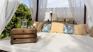Un lit avec une literie blanche, une table de chevet en bois sur laquelle se trouvent une carafe et des verres, et une vue sur la verdure et une clôture.