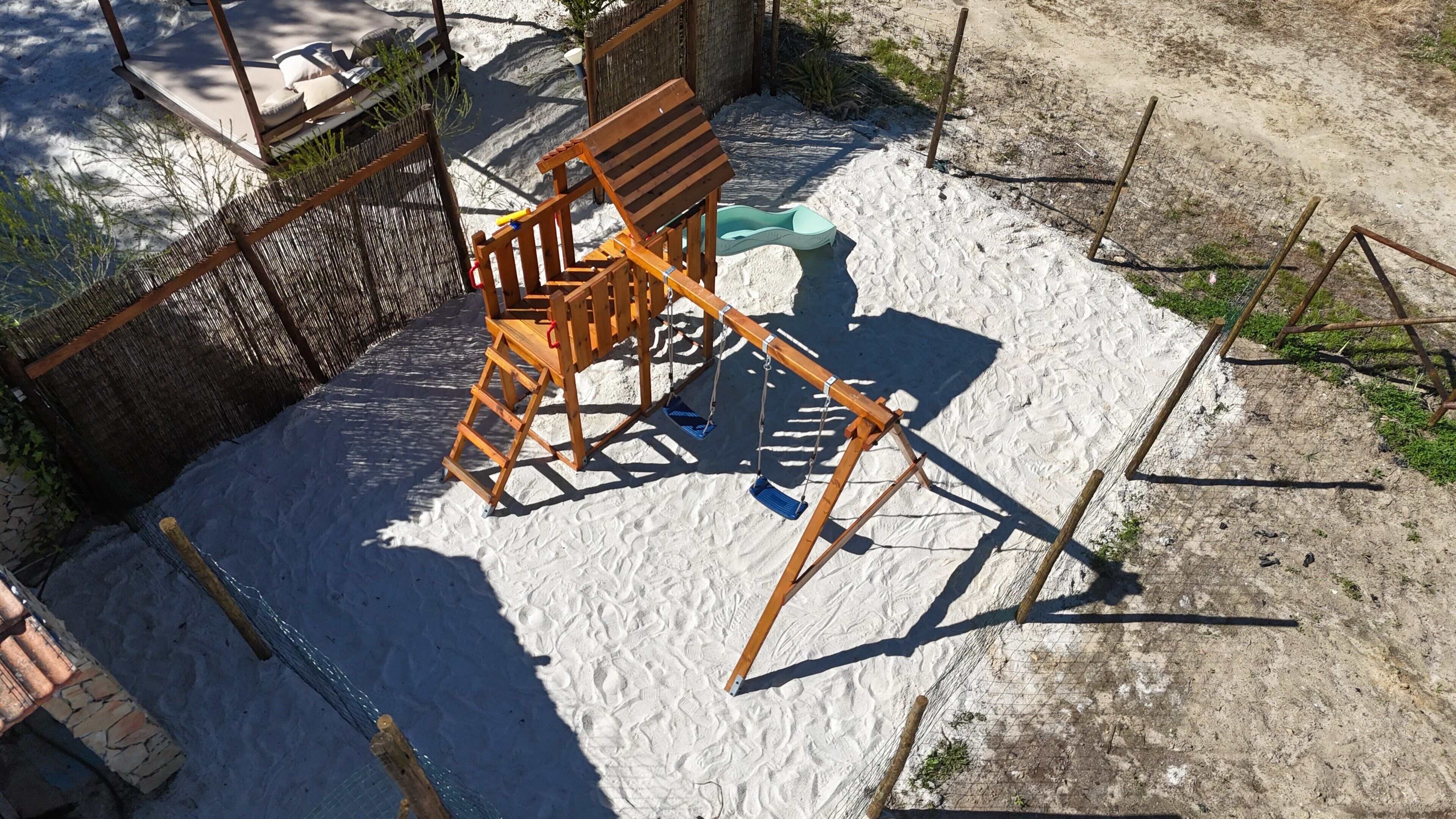 Une balançoire en bois installée dans un espace sablonneux, avec une clôture en fil de fer et un mur en briques.