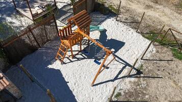 Ein Schaukelstuhl aus Holz in einer Sandfläche mit einem Stahldrahtzaun und einer Backsteinmauer.