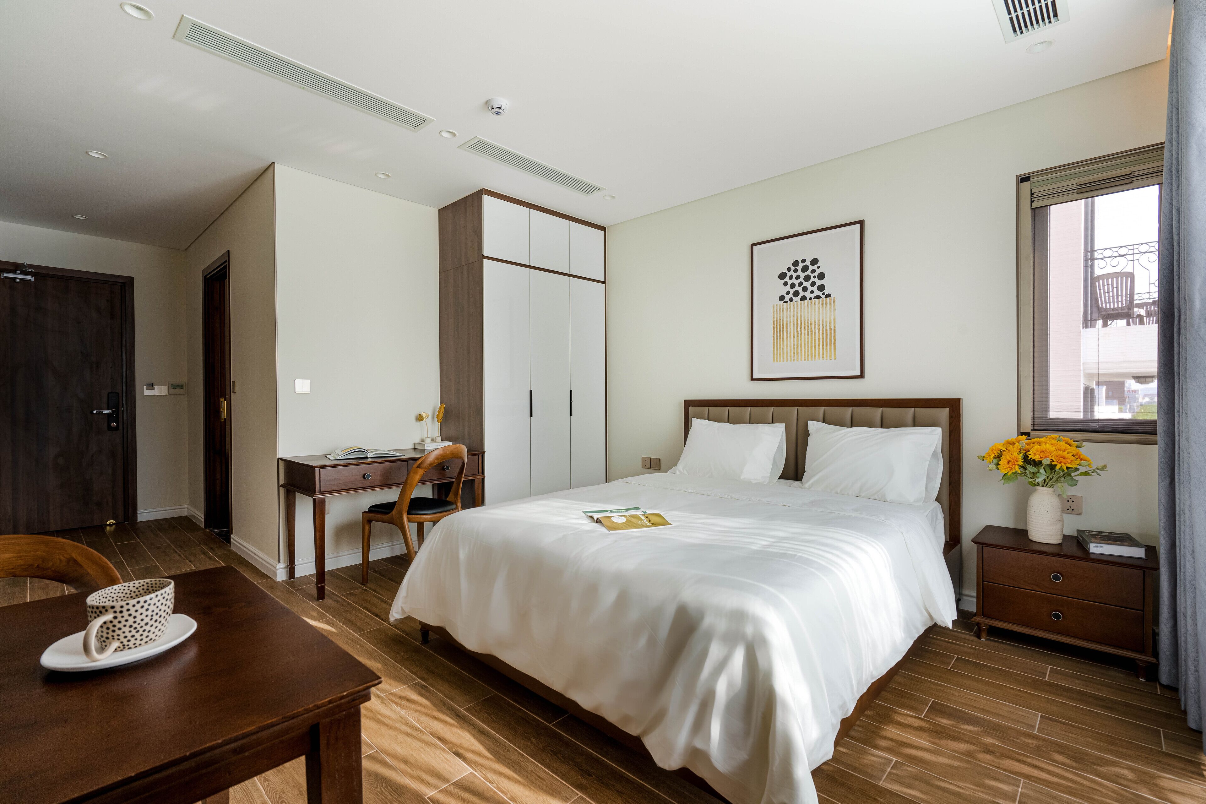 A hotel room with a large bed, a desk, a chair, a small table with a cup, and a window with curtains.