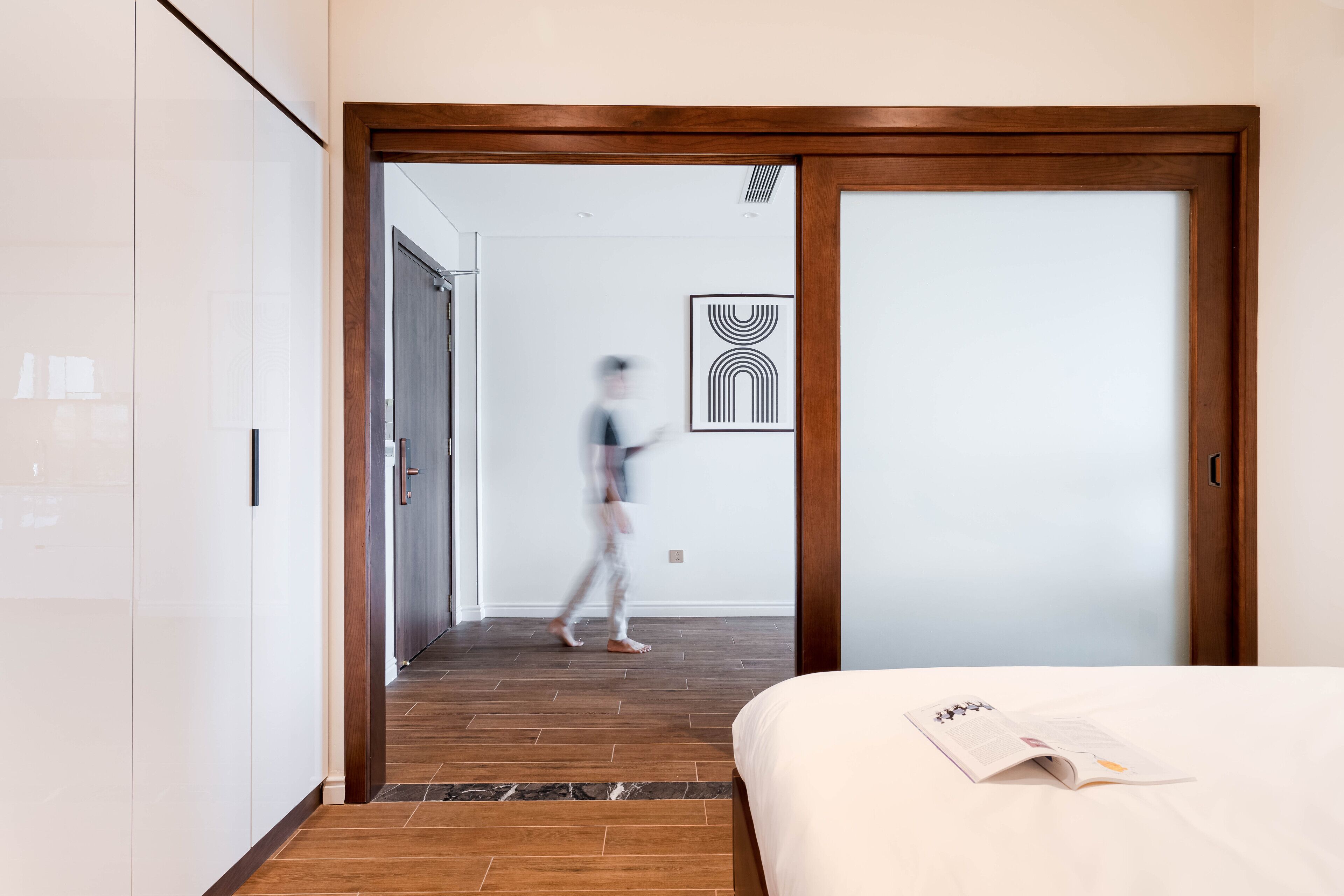A hotel room with a wooden floor, a bed with a magazine on it, and a door leading to another room.