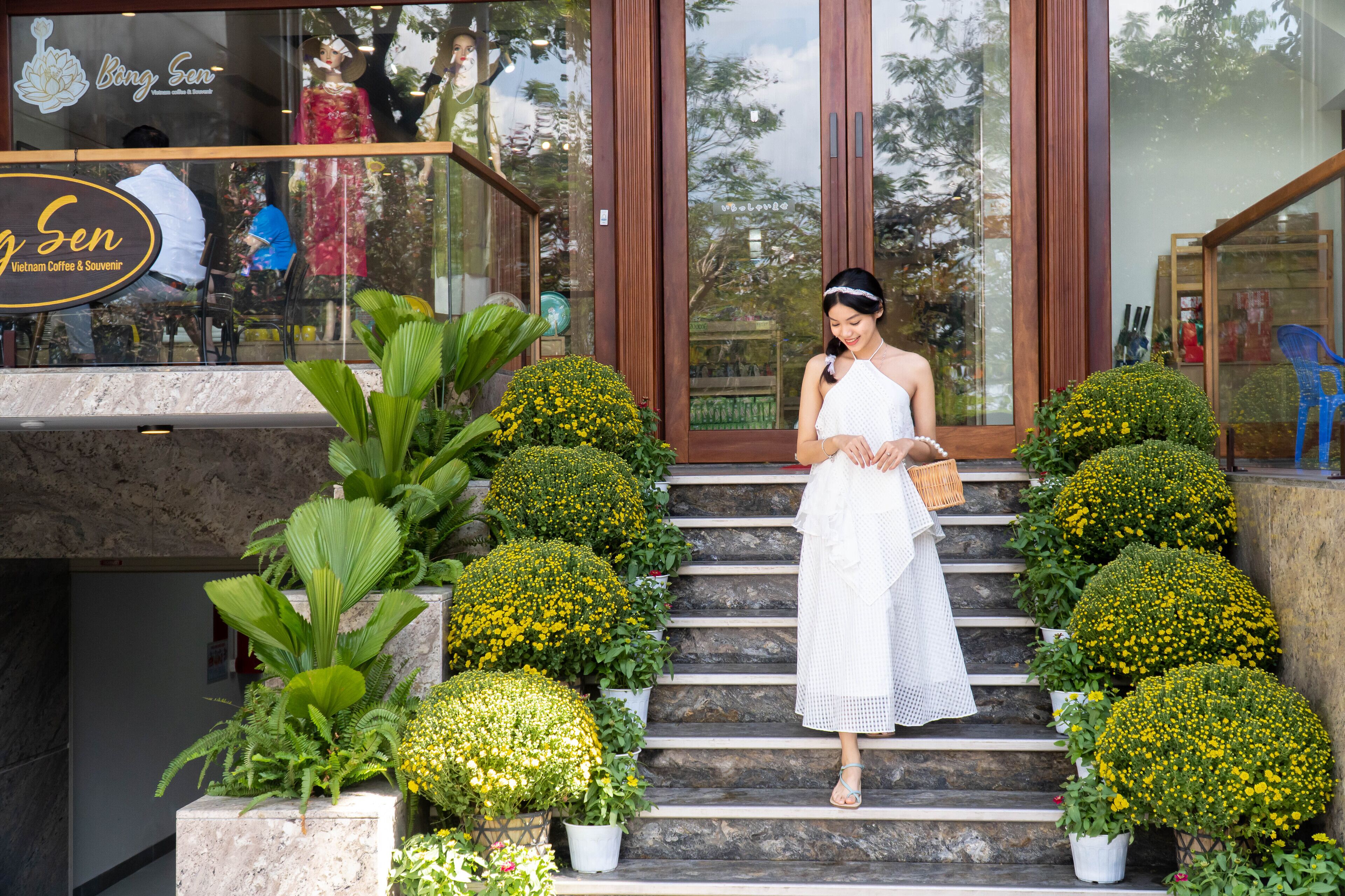 A woman in a white dress standing on stone steps with potted yellow flowers.
