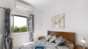 A bedroom with a bed, a nightstand, a lamp, a window with curtains, and a wall-mounted air conditioner. - Casa Verdelho – Retreat Among Vineyards with Lounge, Playground, and Shared Pool (Palmela)
