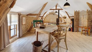 Quarto rústico no sótão com vigas de madeira aparentes, uma mesa de madeira, uma cadeira, um candeeiro pendente e vista pela janela.