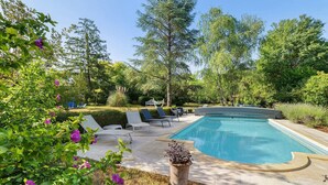 Piscina com espreguiçadeiras, cercada por árvores e gramado.