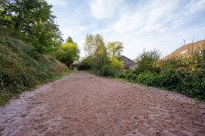 Un chemin de gravier traverse une zone rurale, bordée d’arbres, et un bâtiment se trouve sur la droite.