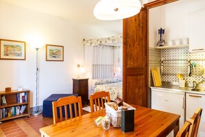 Een kamer met een houten eettafel, stoelen, een boekenkast, een bed met een bloemenbedekking, een nachtkastje met een lamp, en een keuken met een koelkast en planken.
