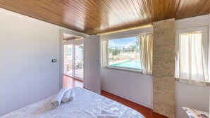 A bedroom with a wooden ceiling, a bed with white linens, a window with curtains, and a view of an outdoor area. - Apartment 'Dependance Con Piscina' with Private Pool, Wi-Fi and Air Conditioning (taurisano)