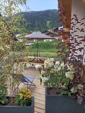 Um deck de madeira com móveis de exterior, rodeado por plantas em vasos e vista para uma montanha.