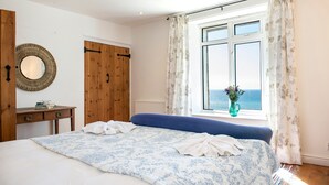 A bedroom with a bed, a wooden wardrobe, a round mirror, a small table, and a window with curtains. - Drake- A Coastal Cottage with Hot Tub & Stunning Views (Portwrinkle)