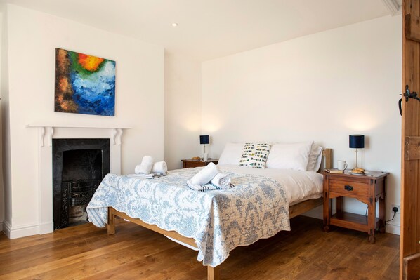 A bedroom with a bed, bedside tables, a fireplace, and a painting on the wall. - Hawkins - A Coastal Cottage with Hot Tub & Stunning Views (Portwrinkle)