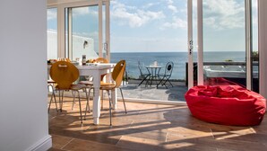 Un comedor con una mesa y sillas, un sillón de tiro rojo y vistas al mar a través de grandes puertas de cristal.
