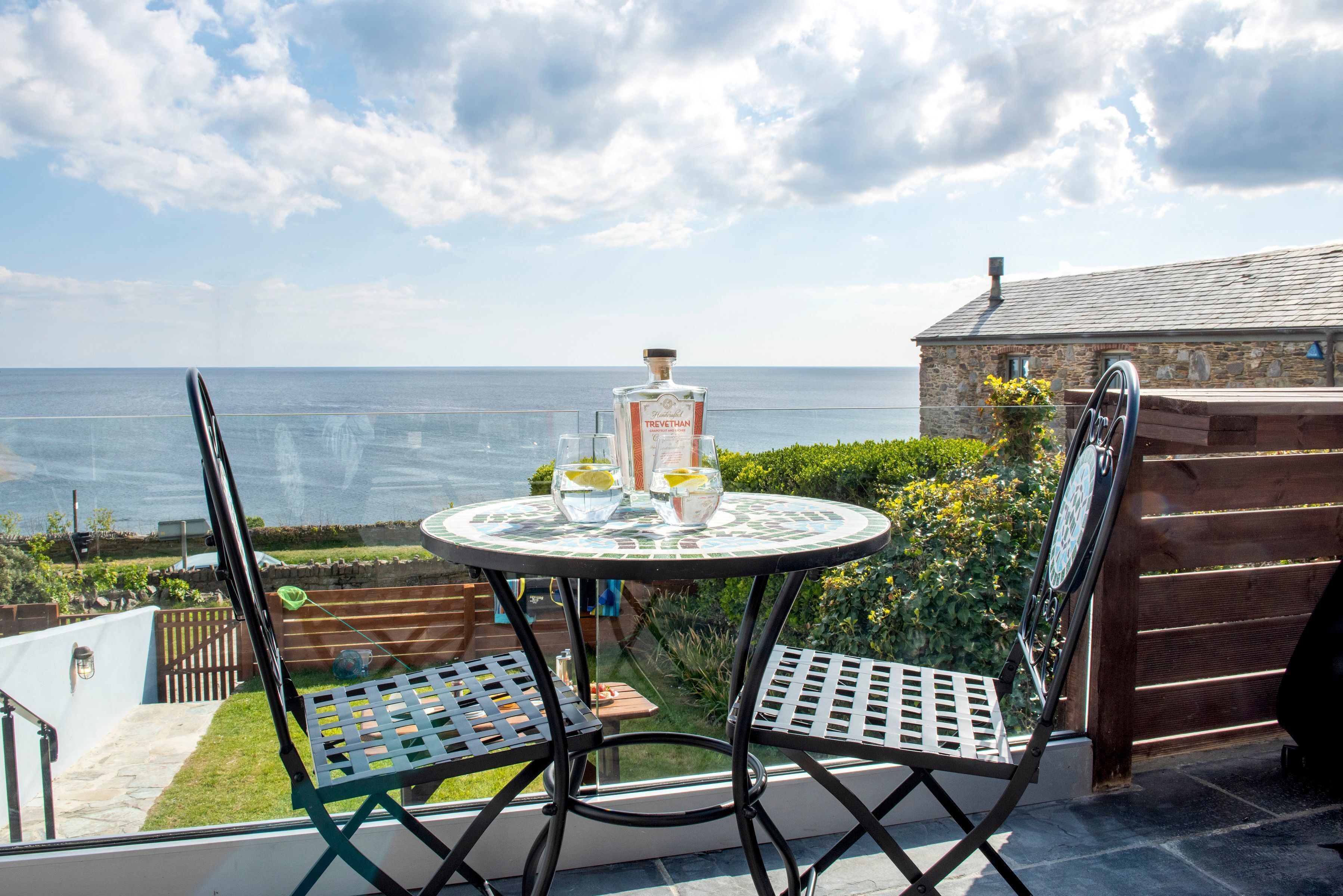 Ein runder Tisch mit zwei Stühlen, einer Flasche Trevethan Gin und Gläsern auf einem Balkon mit Blick auf das Meer.