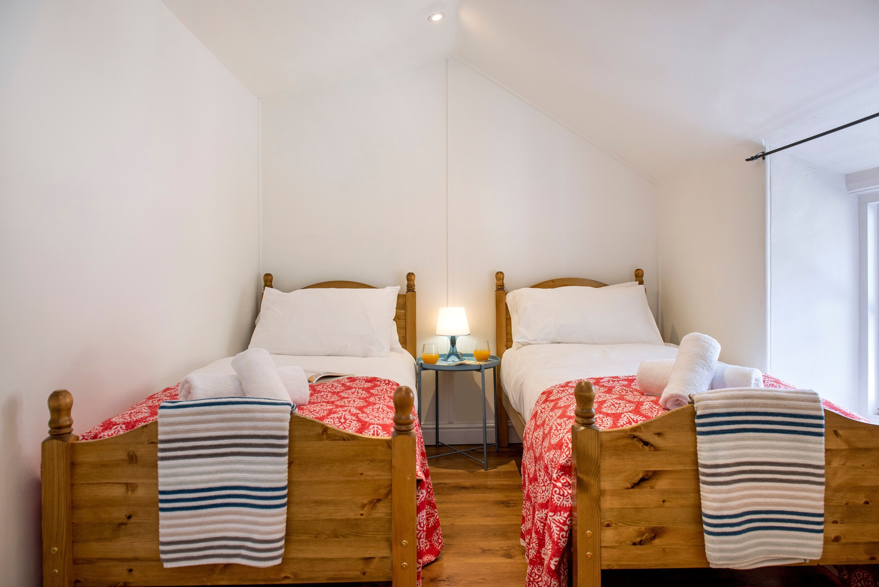Une chambre avec deux lits en bois, chacun recouvert d’une courtepointe à motifs rouges et blancs, avec des oreillers blancs et des serviettes à rayures. Une petite table avec une lampe et deux verres de jus d’orange se trouve entre les lits.