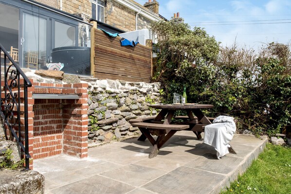 A patio with a brick barbecue, stone steps, a wooden picnic table, and a stone wall. - Grenville - A Coastal Cottage with Hot Tub & Stunning Views (Portwrinkle)