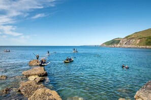 A coastal scene with clear blue water, rocky shore, and people engaging in water activities. - Grenville - A Coastal Cottage with Hot Tub & Stunning Views (Portwrinkle)
