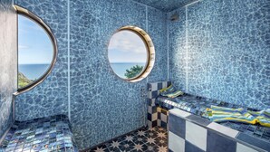 Une salle de bain aux murs recouverts de mosaïques bleues, avec un sol orné de motifs en étoile et une fenêtre circulaire donnant sur la mer.
