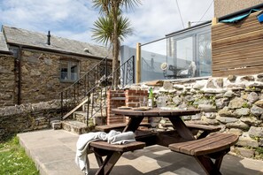 A stone building with a wooden staircase, a brick barbecue area, and a wooden picnic table. - Grenville - A Coastal Cottage with Hot Tub & Stunning Views (Portwrinkle)