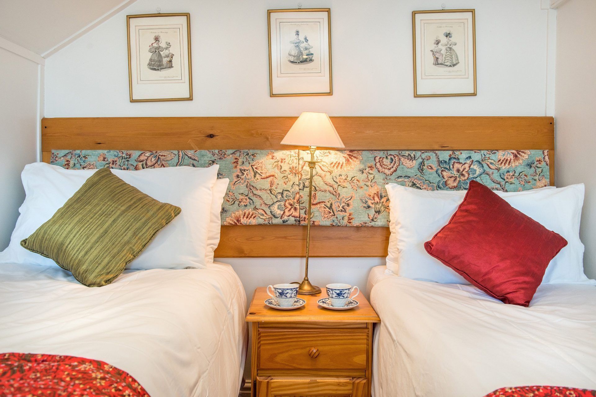 A double bed with a wooden headboard, two pillows, a nightstand with a lamp, and two cups.