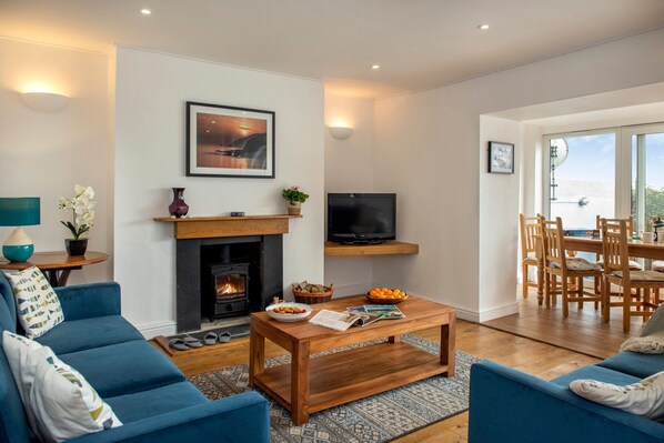 A living room with a fireplace, a television, and a dining area with a view. - Grenville - A Coastal Cottage with Hot Tub & Stunning Views (Portwrinkle)