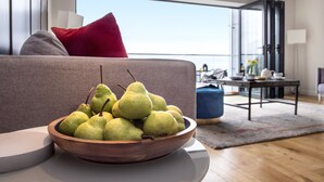 Ein modernes Wohnzimmer mit einer Holtschale voller grüner Birnen auf einem weißen Tisch, einem grauen Sofa mit einem roten Kissen und Blick ins Freie durch große Fenster.