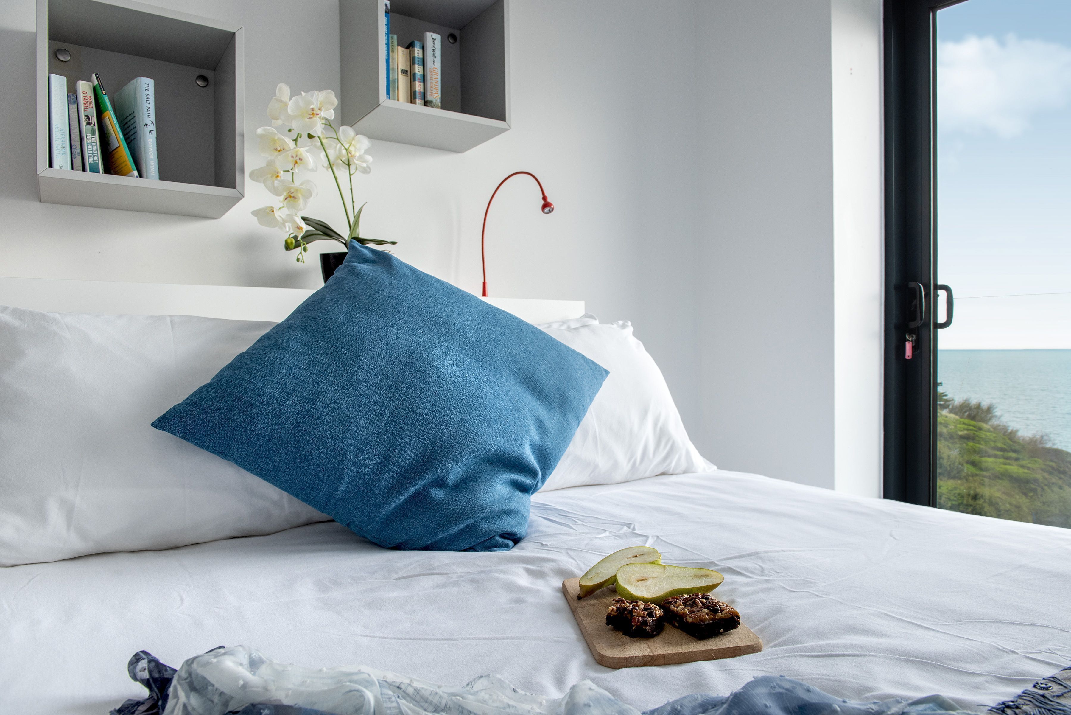 Una cama bien hecha con una almohada azul, una bandeja de madera con aperitivos y un jarrón con flores blancas. Hay estanterías sobre la cama y una vista al mar a través de una ventana.
