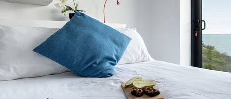 Una cama bien hecha con una almohada azul, una bandeja de madera con aperitivos y un jarrón con flores blancas. Hay estanterías sobre la cama y una vista al mar a través de una ventana.