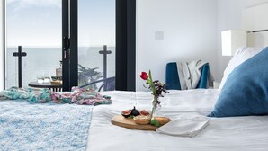 Ein Hotelzimmer mit Bett, einem kleinen Tisch mit einer Essschale, einer Vase mit einer Blume und Meerblick durch große Fenster.