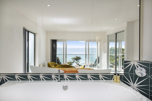 A modern bathroom with a large bathtub, a view of the sea, and a balcony with seating.