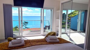 A hotel room with a bed, two towels, a patterned rug, and a view of the sea and greenery through large glass doors. - Turnstone -  Beach-side House with Terrace Hot Tub & Stunning Views (Portwrinkle)