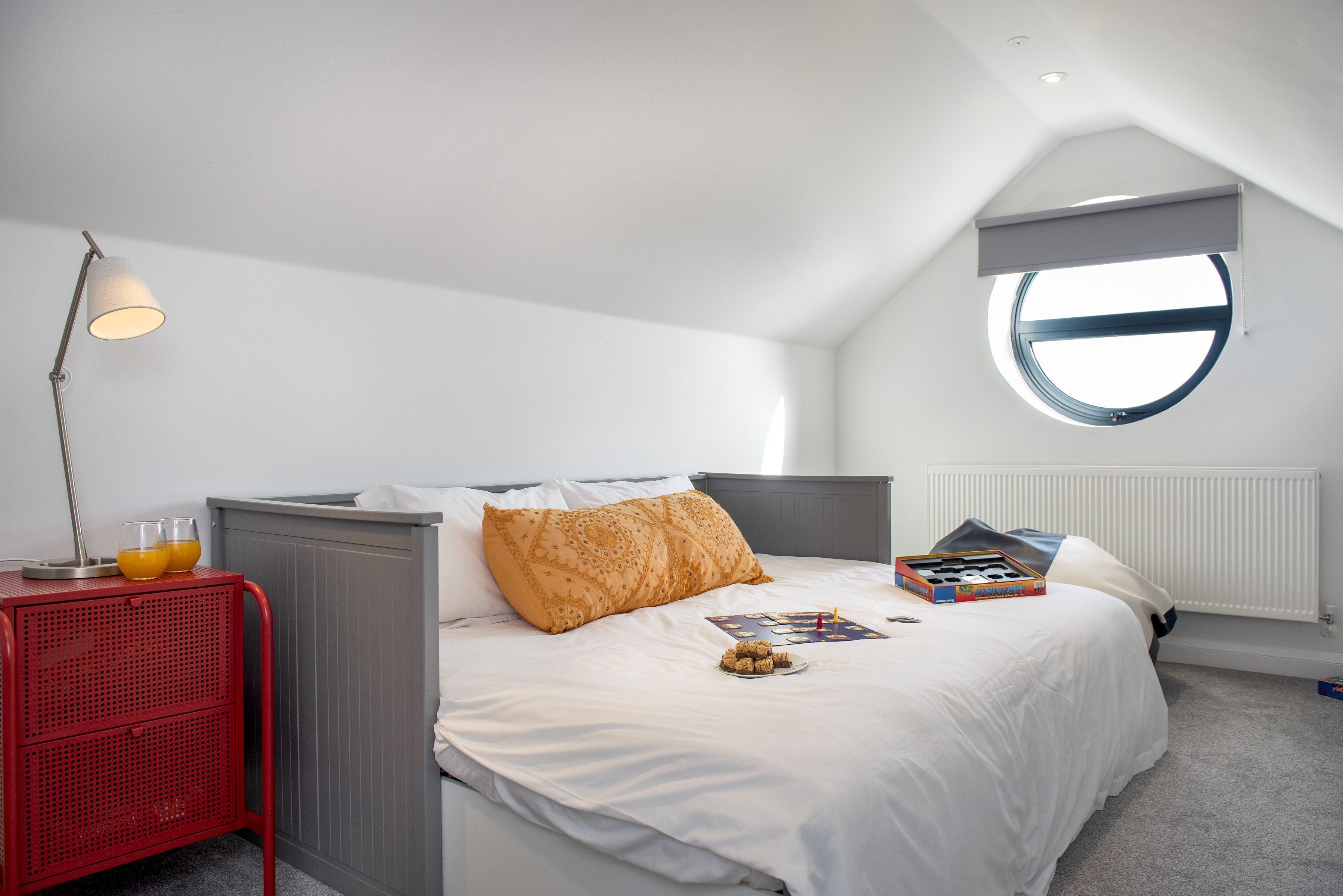 A bedroom with a bed, a red side table, a lamp, a round window, and a radiator.