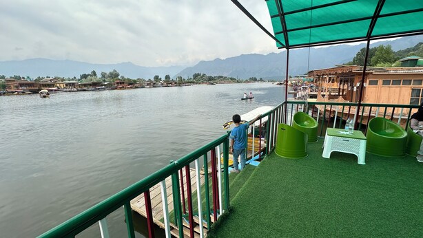 A river view from a covered deck with a green canopy, wooden boats, and a distant mountain range. - The Heritage Safina Group Of Houseboats (Srinagar)