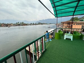 Vista de un río desde un porche cubierto con toldo verde, botes de madera y una cadena montañosa distante.