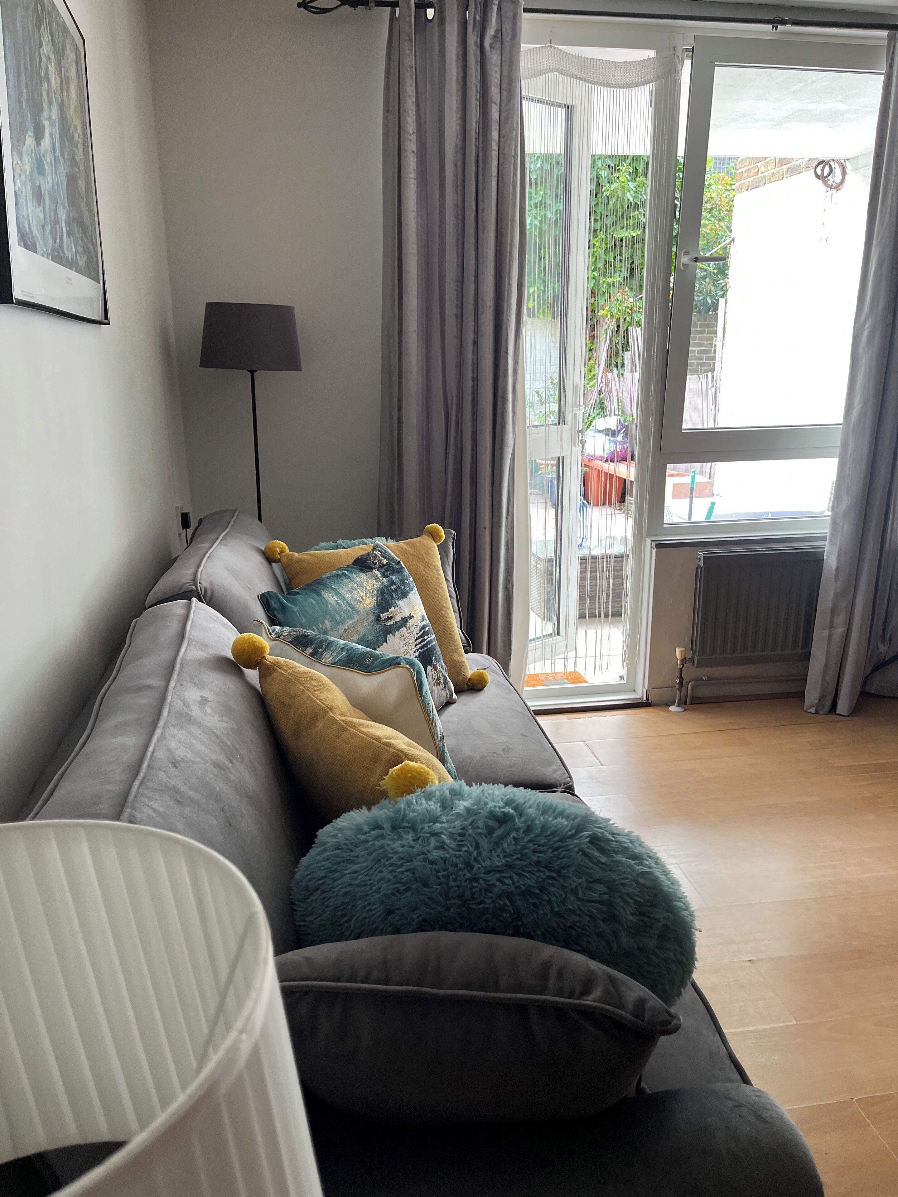 A modern living room with a grey sofa, multiple cushions, a lamp, and a view of the outdoors through a sliding glass door.