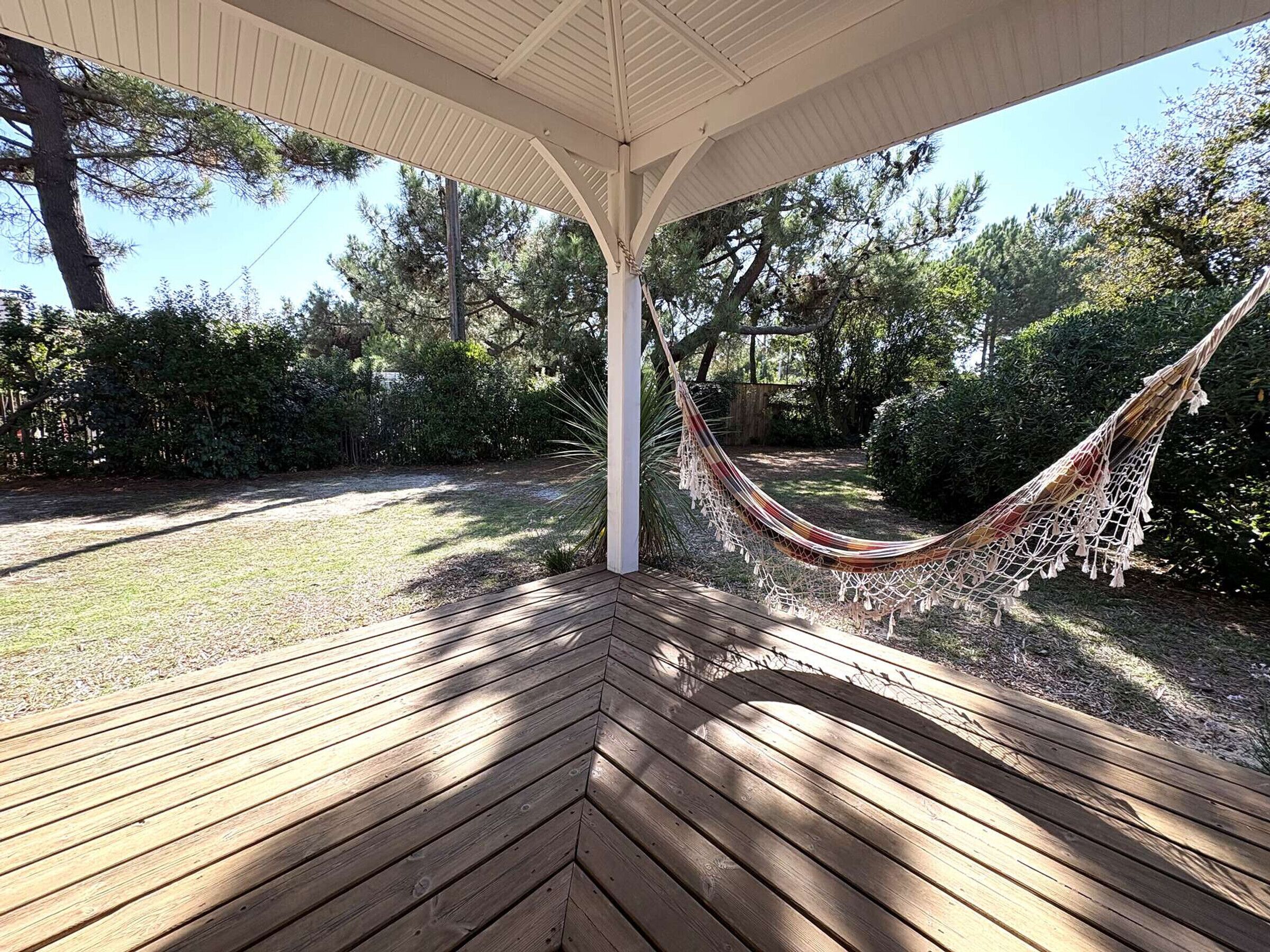 A wooden deck with a hammock under a white pergola, surrounded by greenery.