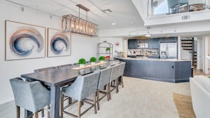 Une cuisine moderne dotée d’un îlot central, un coin repas avec une longue table et des chaises, et une chambre en mezzanine avec un balcon.