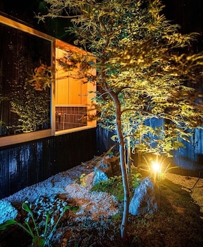 A modern building with a wooden facade and a well-lit garden area featuring a tree and rocks.