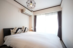 A hotel room with a large bed, a nightstand with a lamp, an air conditioning unit, and a window with curtains.